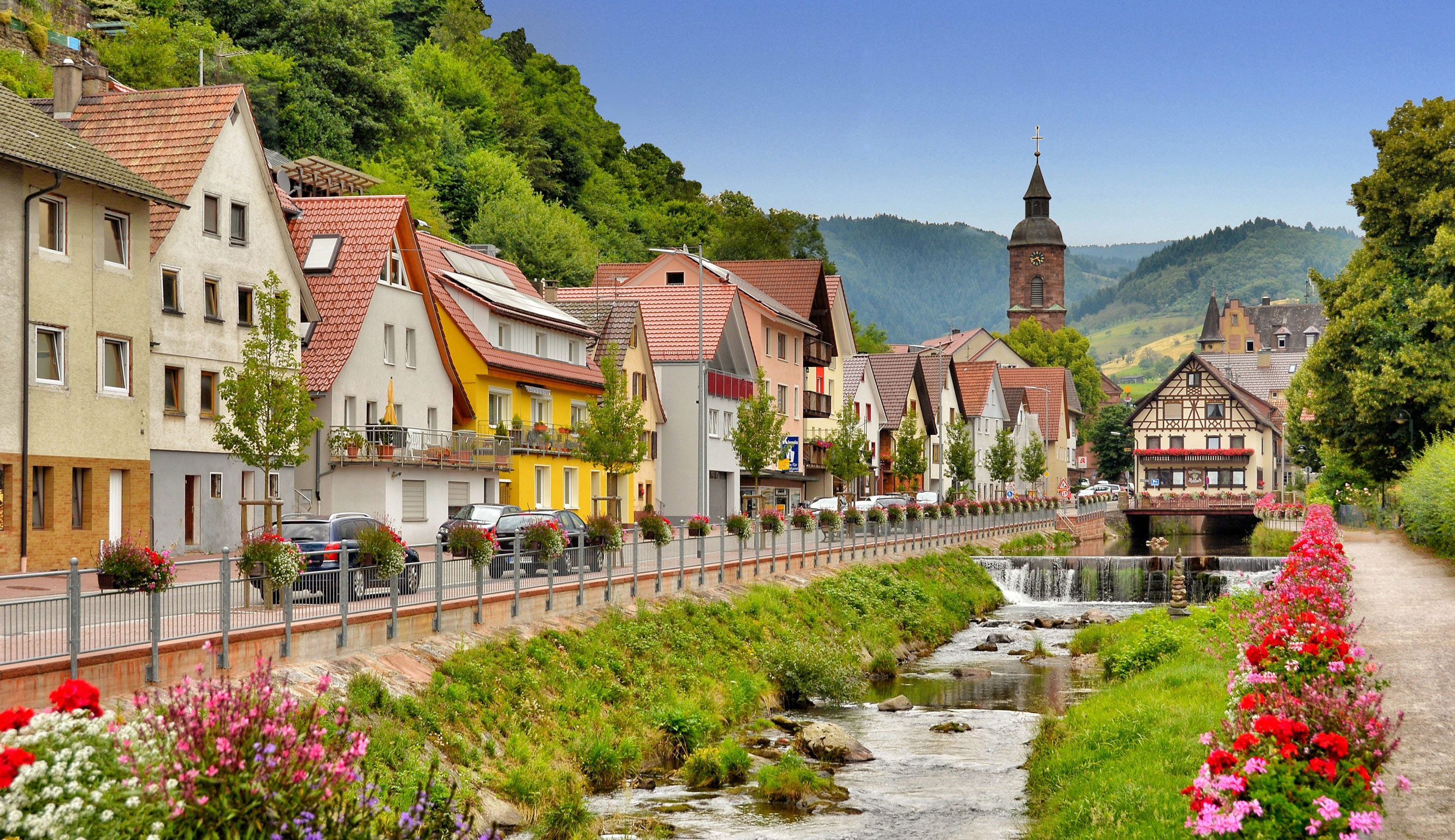Häuserreihe entlang eines Baches. Ufer bepflanzt mit Blumen. Im Hintergrund Berge